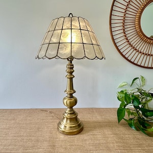 May include: A table lamp with a brass-toned metal base and a scalloped, translucent white shade featuring a grid pattern. The lamp has a tiered, rounded stem and a circular base, emitting a warm light from within the textured, panelled shade.