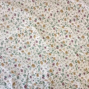May include: White fabric with a floral pattern. Small, light purple and yellow flowers are scattered across the material, with green leaves and stems. The fabric has a slightly creased texture.