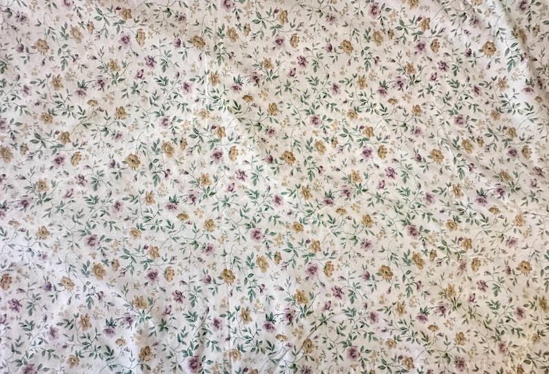 May include: White fabric with a floral pattern. Small, light purple and yellow flowers are scattered across the material, with green leaves and stems. The fabric has a slightly creased texture.