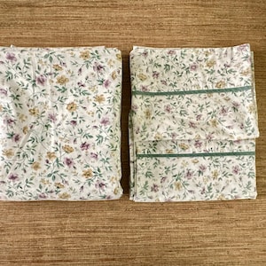 May include: Two folded floral bed sheets. The sheets have a white background with a repeating pattern of purple, yellow, and green flowers and leaves. One sheet has green trim.