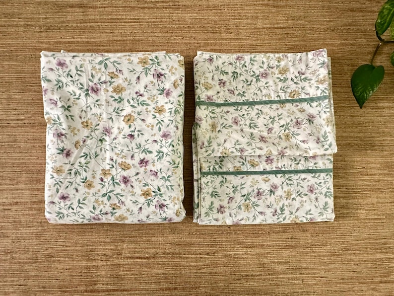 May include: Two folded floral bed sheets. The sheets have a white background with a repeating pattern of purple, yellow, and green flowers and leaves. One sheet has green trim.