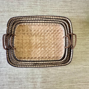 May include: A set of three rectangular woven baskets nested together. Each basket features a light tan woven base with a darker brown woven rim and two matching brown handles on the sides. The baskets are arranged in a stack, showing the interior.