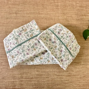 May include: Two folded pillowcases with a floral pattern of purple, yellow, and green flowers on a white background. Each pillowcase has a green border. The pillowcases are on a wooden surface.