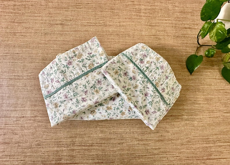 May include: Two folded pillowcases with a floral pattern of purple, yellow, and green flowers on a white background. Each pillowcase has a green border. The pillowcases are on a wooden surface.