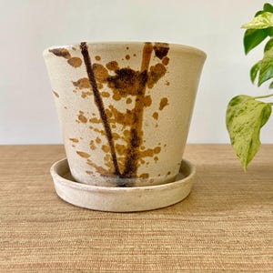 May include: A tapered ceramic plant pot with a matching circular saucer. The pot features a light cream-colored base with dark brown, irregular paint-like splatters and vertical streaks that extend from the rim toward the base of the vessel.