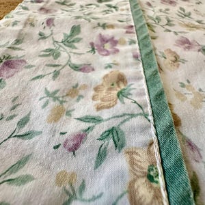 May include: Close-up of a floral fabric with a white background. The fabric features a pattern of purple, yellow, and green flowers and leaves. A green border runs along the edge of the fabric, adding a contrasting detail. The fabric appears to be cotton or linen.