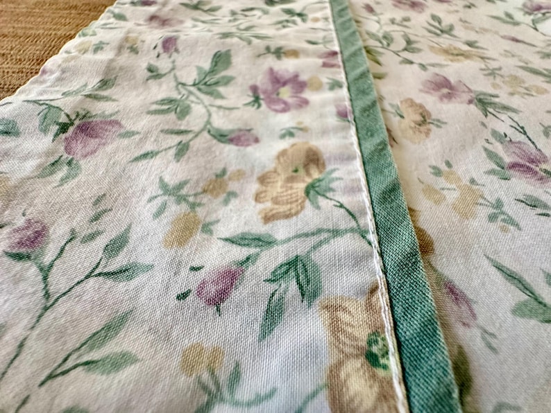 May include: Close-up of a floral fabric with a white background. The fabric features a pattern of purple, yellow, and green flowers and leaves. A green border runs along the edge of the fabric, adding a contrasting detail. The fabric appears to be cotton or linen.