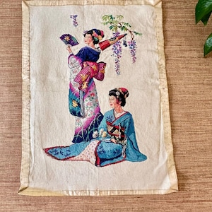 May include: A rectangular fabric wall hanging featuring an embroidered scene of two figures in traditional attire. One figure stands holding a purple fan, while the other sits below. The design includes wisteria branches and floral patterns in shades of blue, pink, and purple on an off-white background with a light-colored fabric border.