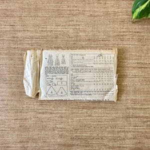 May include: A vintage sewing pattern packet, featuring illustrations of dress designs and detailed instructions. The packet is off-white with visible wear and tear, and includes text and diagrams. The background is a textured, light brown surface.