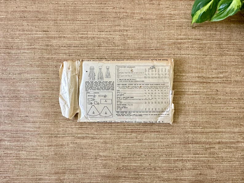 May include: A vintage sewing pattern packet, featuring illustrations of dress designs and detailed instructions. The packet is off-white with visible wear and tear, and includes text and diagrams. The background is a textured, light brown surface.
