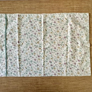 May include: A rectangular white floral fabric featuring a pattern of small purple, yellow, and green flowers and leaves. A light blue trim runs along the left edge. The fabric is likely a tablecloth or similar textile.