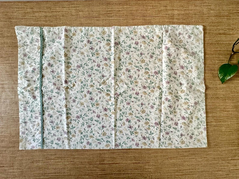 May include: A rectangular white floral fabric featuring a pattern of small purple, yellow, and green flowers and leaves. A light blue trim runs along the left edge. The fabric is likely a tablecloth or similar textile.