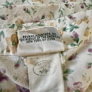 May include: Close-up of a floral fabric with a cream background and a pattern of purple, green, and tan flowers and leaves. The fabric has a tag with the text "REVMAN INDUSTRIES, INC. 1211 AVENUE OF THE AMERICAS NEW YORK, NY 10036".