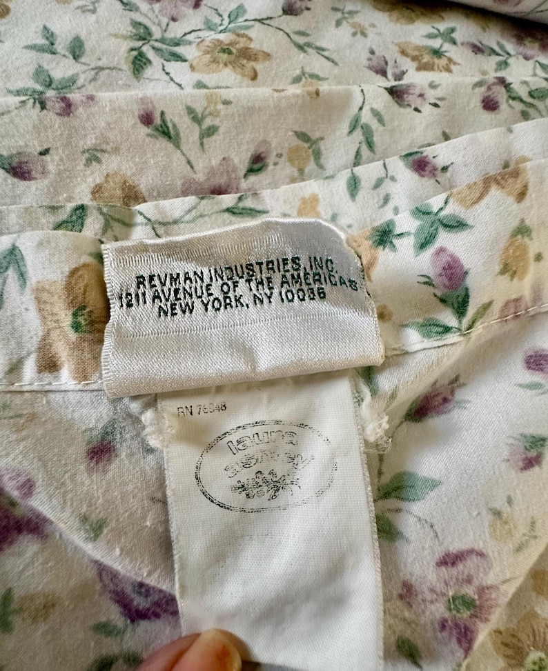 May include: Close-up of a floral fabric with a cream background and a pattern of purple, green, and tan flowers and leaves. The fabric has a tag with the text "REVMAN INDUSTRIES, INC. 1211 AVENUE OF THE AMERICAS NEW YORK, NY 10036".