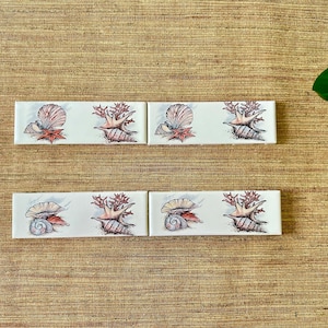 May include: Six white ceramic tiles with a seashell and coral design. Each tile features a different seashell and coral arrangement.