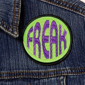 May include: A green and purple embroidered patch with the word "FREAK" in a circle. The patch is sewn onto a blue denim jacket.