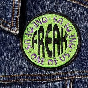 Könnte beinhalten: Ein gestickter Aufnäher mit dem Text "One of Us, One of Us, Freak" in einem Kreis, mit grüner und violetter Stickerei.