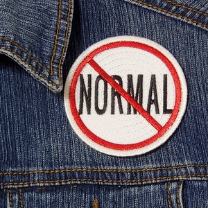 Puede incluir: Chaqueta vaquera azul con un parche redondo. El parche muestra la palabra "NORMAL" en negro, tachada con una línea roja. El parche tiene un fondo blanco y un borde rojo. La chaqueta tiene botones plateados.