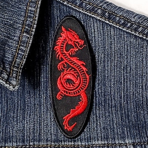 May include: Red embroidered dragon patch on a denim jacket. The dragon is curled up in a circle with its head facing the right. The patch is oval shaped and has a black background.