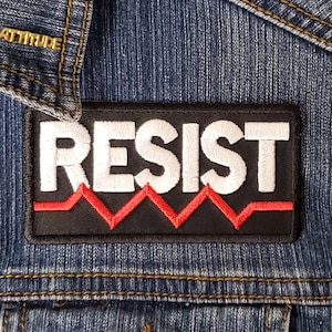 May include: A black and red embroidered patch with the word "RESIST" in white letters. The patch is sewn onto a blue denim jacket.