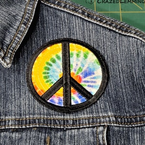 May include: A black peace sign patch with a tie-dye design in yellow, orange, red, blue, and green colors. The patch is sewn onto a denim jacket.