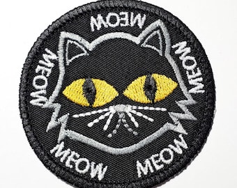 Op de afbeelding: Een zwarte ronde patch met een kattengezichtontwerp. De kat heeft gele ogen, witte snorharen en het woord "MEOW" is in het wit rond de rand geborduurd. De patch is gemaakt van stof.