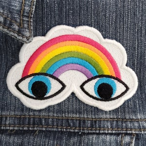 May include: A white cloud with a rainbow and two large eyes embroidered on a denim jacket. The rainbow is made of red, orange, yellow, green, blue, indigo, and violet thread.