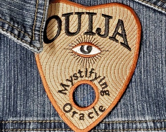 May include: A brown felt patch with the word "OUIJA" in black embroidery. The patch has a stylized eye with a black pupil and a circle in the center. The text "Mystifying Oracle" is embroidered around the circle.
