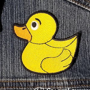 Puede incluir: Un pato de goma amarillo con contorno negro, bordado en una tela vaquera azul.