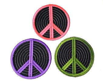Könnte beinhalten: Drei gestickte Friedenssymbol-Patches in Pink, Lila und Grün. Jeder Patch hat einen schwarzen Hintergrund mit konzentrischen Kreisen und einem weißen Umriss.