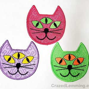 May include: Three embroidered iron-on patches featuring a cartoon cat with three eyes. The cat is smiling and has a pink, purple, and green face.