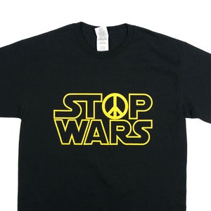Op de afbeelding: Zwart T-shirt met een gele afbeelding met de tekst "STOP WARS" met een vredesteken in het midden van het woord "STOP".