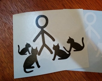May include: A white sticker featuring a black stick figure surrounded by four black cat silhouettes. A second sticker with a similar design is partially visible in the background. The design is simple and graphic, suitable for cat lovers.
