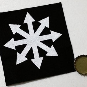 Op de afbeelding: Een zwarte vierkante patch met een wit achtpuntig sterburst-ontwerp met pijlen die naar buiten wijzen. Een goudkleurige flesdop ligt naast de patch.