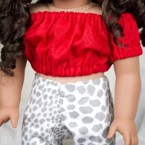 May include: A doll's outfit featuring a red, off-the-shoulder top and patterned trousers. The top has short sleeves and a gathered design. The trousers are patterned with grey spots on a white background. A fashionable doll's ensemble.