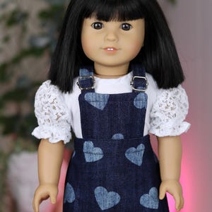 May include: A doll with black hair and brown eyes wearing a white lace-sleeved shirt and a denim jumper. The jumper features a pattern of blue hearts and is secured with silver buckles. The doll is a child's toy.