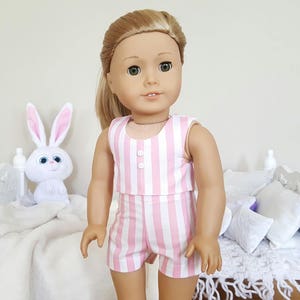 May include: A pink and white striped sleeveless jumpsuit with a button closure. The jumpsuit is made for a doll and is being worn by a doll with blonde hair.