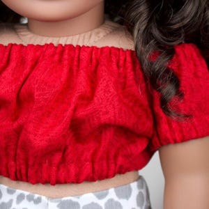 May include: A red off-the-shoulder top with short sleeves. The top is made of a textured fabric and is paired with a light-coloured garment with a grey and white pattern. Dark brown hair is visible on the right side.