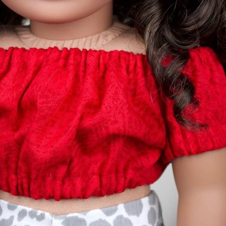 May include: A red off-the-shoulder top with short sleeves. The top is made of a textured fabric and is paired with a light-coloured garment with a grey and white pattern. Dark brown hair is visible on the right side.
