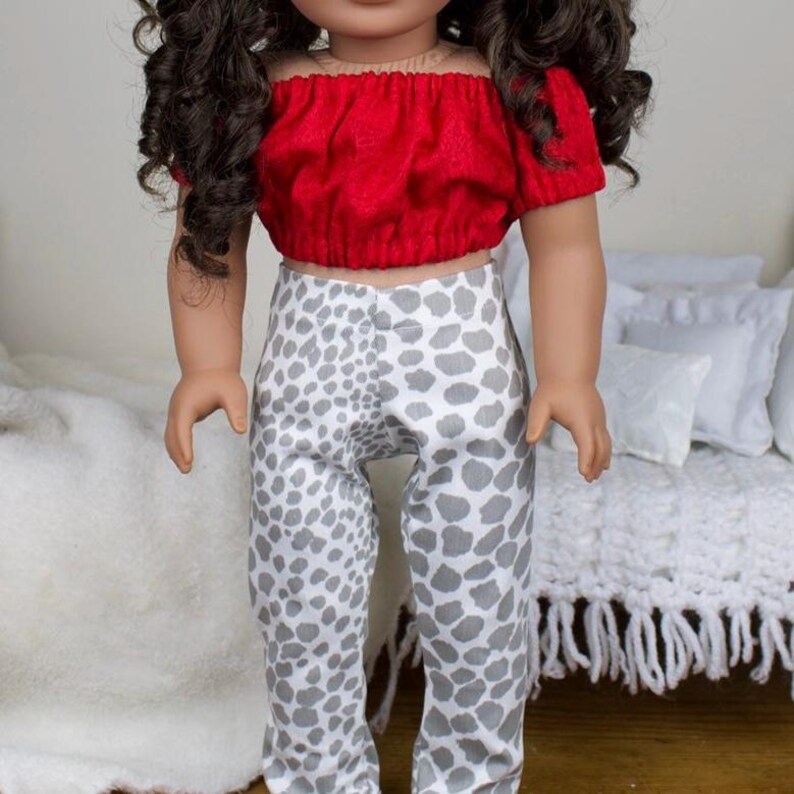 May include: A doll dressed in a red, off-the-shoulder crop top and grey and white animal print trousers. The doll has curly brown hair. The outfit is designed for a doll.