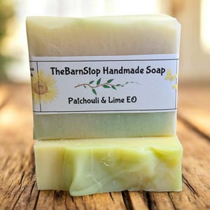 Pode incluir: Uma barra retangular de sabonete artesanal verde-claro embrulhada num rótulo de papel branco. O rótulo apresenta o texto TheBarnStop Handmade Soap e Patchouli & Lime EO, decorado com uma ilustração de girassol amarelo e um pequeno ramo.