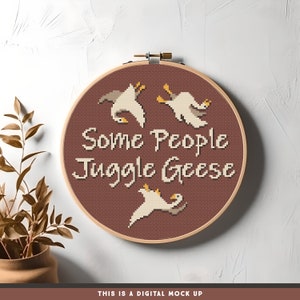 May include: A brown embroidery hoop with a white background and the text "Some People Juggle Geese" in white. Three white geese are embroidered in the design, two are upside down and one is right side up.