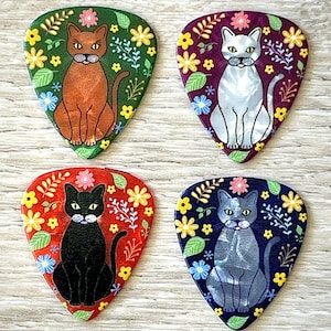 May include: Four guitar picks with cat illustrations. Each pick features a different cat color: brown, gray, black, and blue. The cats are surrounded by floral designs in various colors, including yellow, pink, and blue. The picks are on a light wood surface.