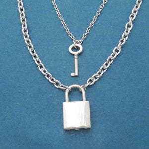 Puede incluir: Conjunto de collares plateados con un colgante de candado en una cadena gruesa, con un colgante de llave más pequeño en una cadena más fina encima. Se muestra sobre un fondo azul liso.