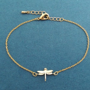 Könnte beinhalten: Ein goldenes Kettenarmband mit einem Libellen-Anhänger. Die Kette ist zart und die Libelle detailliert.