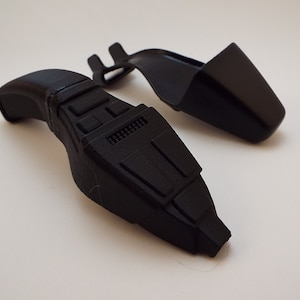 May include: A black plastic 3D printed handheld device with a textured surface and a handle. The device has a series of buttons and a small screen. The device is accompanied by a separate black plastic 3D printed piece.