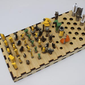 May include: A rectangular wooden storage rack with a grid of circular holes, holding a collection of various metal router bits. The bits feature diverse cutting profiles and include yellow, green, orange, and silver-coloured components.