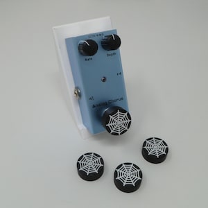 May include: A light blue analog chorus pedal with black knobs and a spiderweb design on the knobs. The pedal is mounted on a white plastic base.