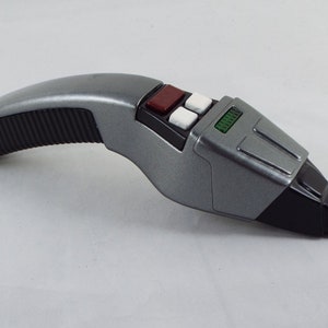 Könnte beinhalten: Ein silbernes und schwarzes Star Trek Phaser-Spielzeug mit einem roten und weißen Knopfpanel. Der Phaser hat einen strukturierten schwarzen Griff und ein grünes digitales Display.