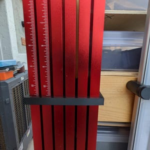 Puede incluir: Una herramienta de medición de metal rojo con barras verticales y una base negra. La herramienta tiene una regla impresa en una de las barras, con medidas en centímetros. Diseñada para carpintería u otras tareas de precisión.